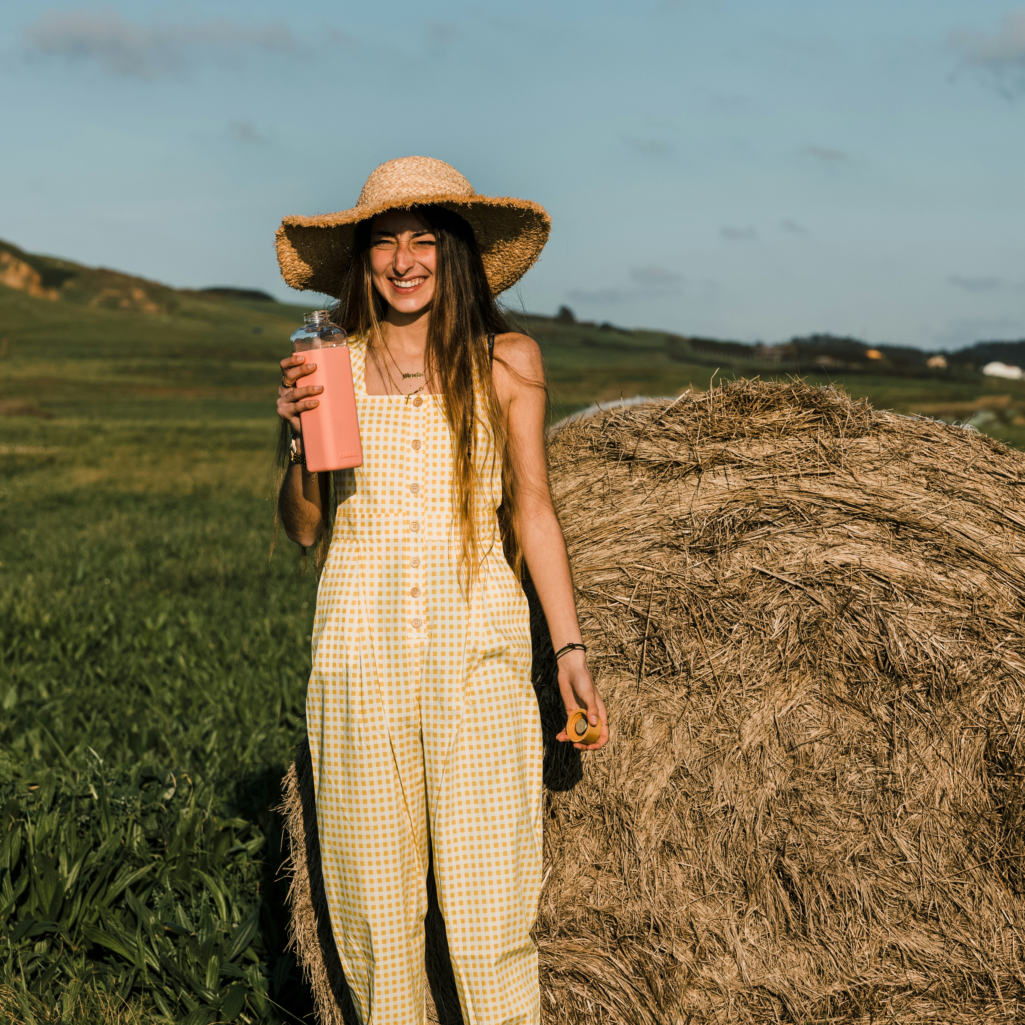 woman in brown and white plaid dress wearing brown straw hat standing on brown grass field